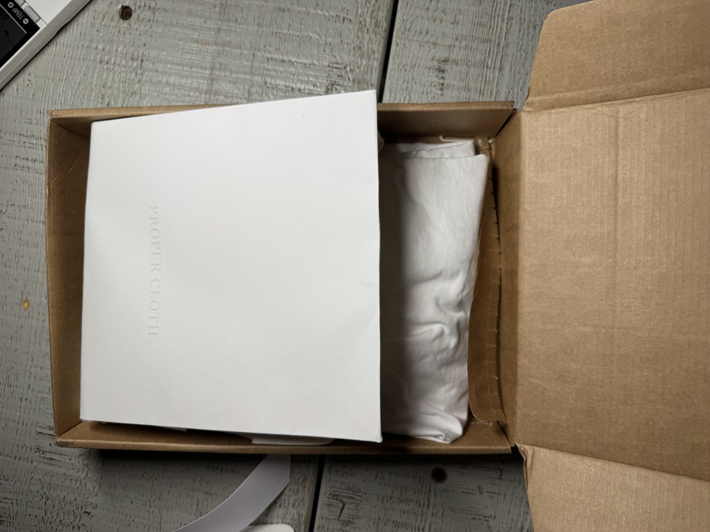An opened Proper Cloth shipping box with the folded shirt visible inside, in kraft-paper packaging
